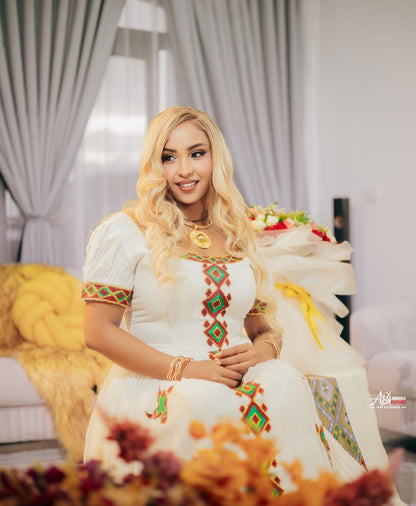 A woman with long blonde wavy hair is seated indoors wearing a white traditional habesha kemis with colorful geometric embroidery in green, red, and yellow. She is posed in a bright room with gray curtains, a yellow chair, and floral arrangements in the background.