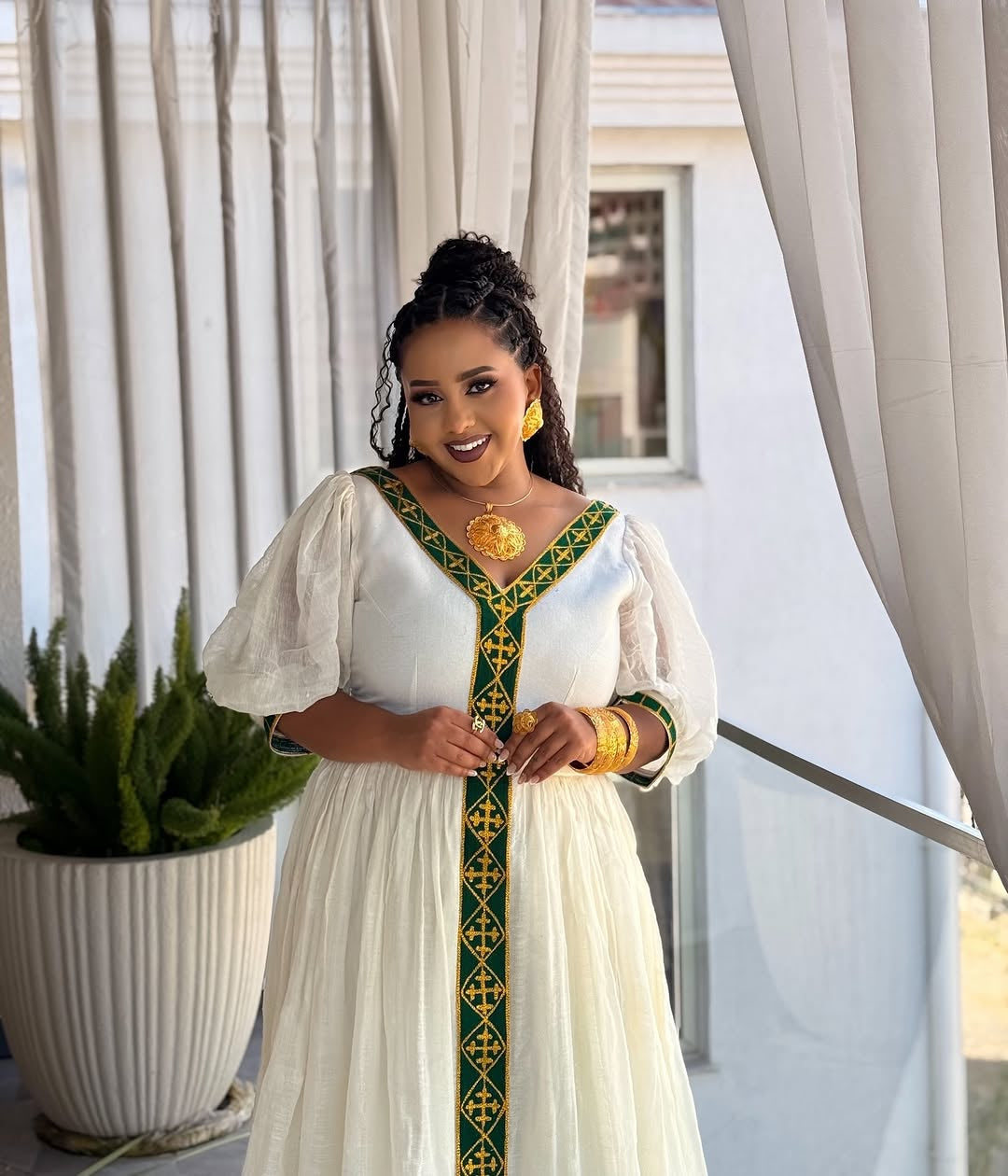 Woman in a white dress with green and gold  Ethiopian dress standing in front of white curtains.