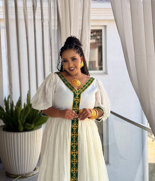 Woman in a white dress with green and gold  Ethiopian dress standing in front of white curtains.