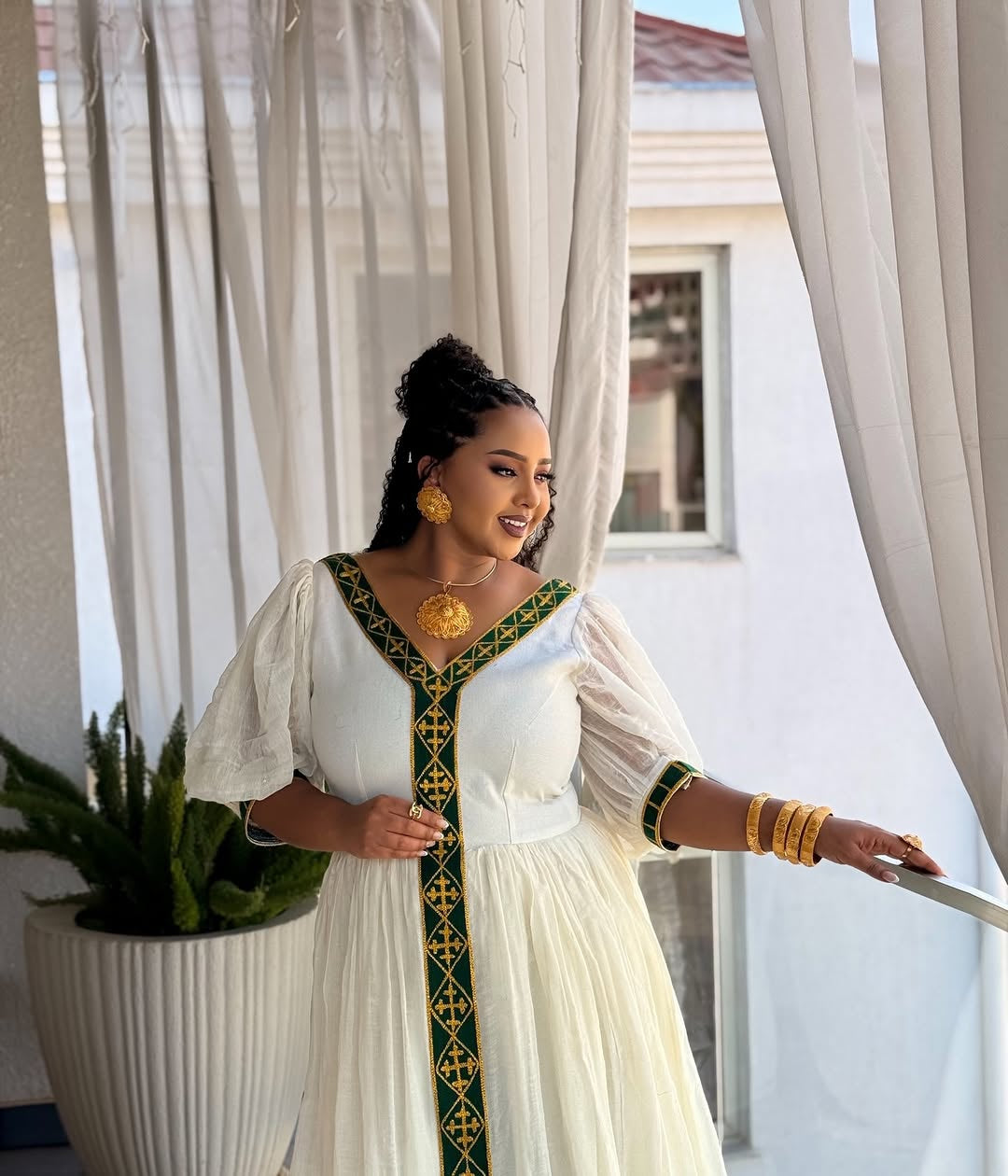 Woman in a white dress with green patterns Ethiopian dress standing in a bright room with white curtains.