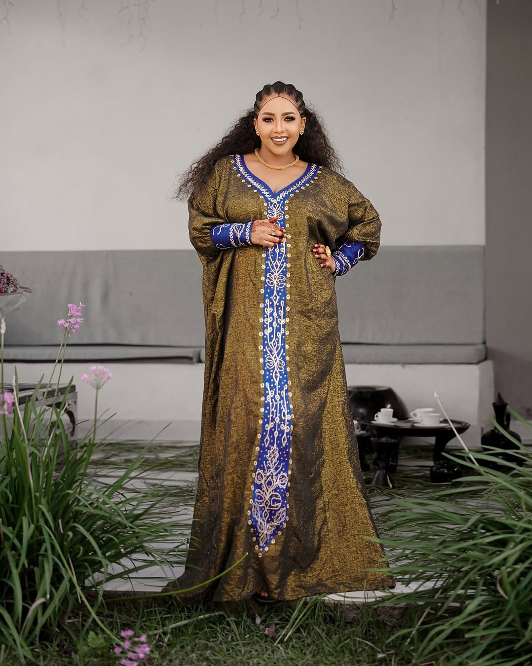 Woman in a long, patterned Habesha Ethiopian Dress standing outdoors with greenery and a table in the background.