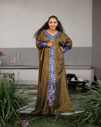 Woman in a long, patterned Habesha Ethiopian Dress standing outdoors with greenery and a table in the background.