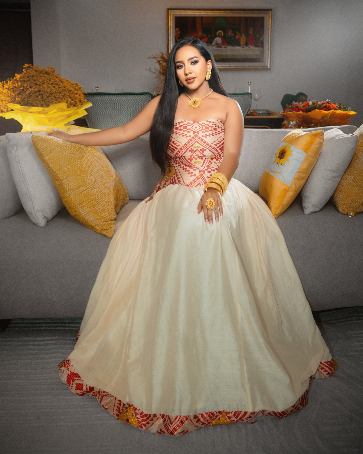 A woman wearing a voluminous off-white ball Ethiopian National Dresses with a red and gold patterned bodice and hemline, seated on a gray sofa with decorative pillows in a living room setting.