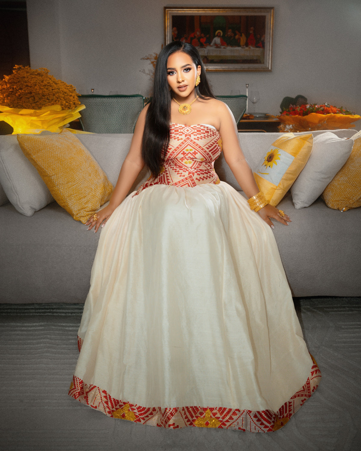 A woman wearing a strapless ball Ethiopian National Dresses with a cream-colored skirt and a red and gold embroidered bodice, sitting on a gray sofa with yellow and white pillows in a living room setting.