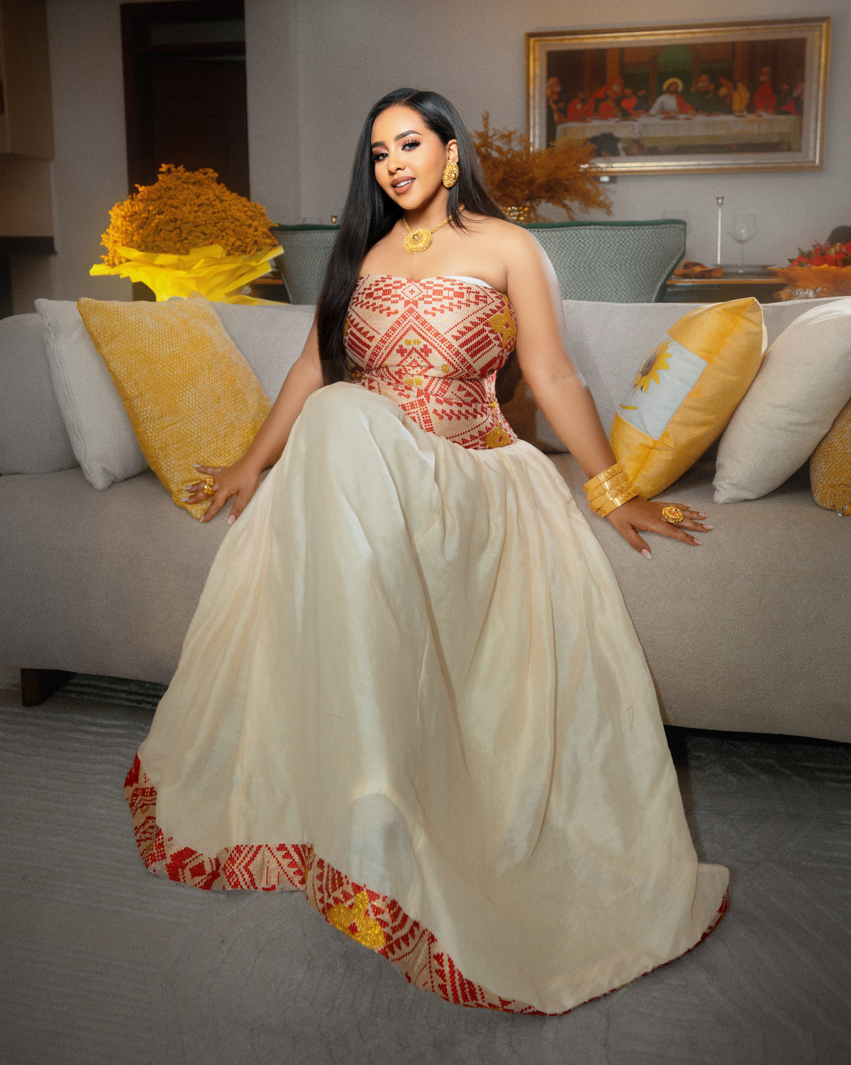 A woman wearing a strapless formal Ethiopian National Dresses with a red and gold geometric embroidered bodice and a voluminous cream skirt, seated on a gray sofa with yellow pillows in a decorated living room.