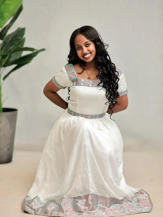 A woman wearing a white habesha kemis with a patterned border and short sleeves, kneeling on a light floor while a man adjusts the hem of the dress.