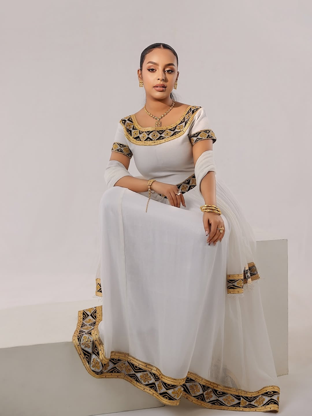 Woman wearing a white saree with black and gold Habesha Zuria embroidery sitting on a white background