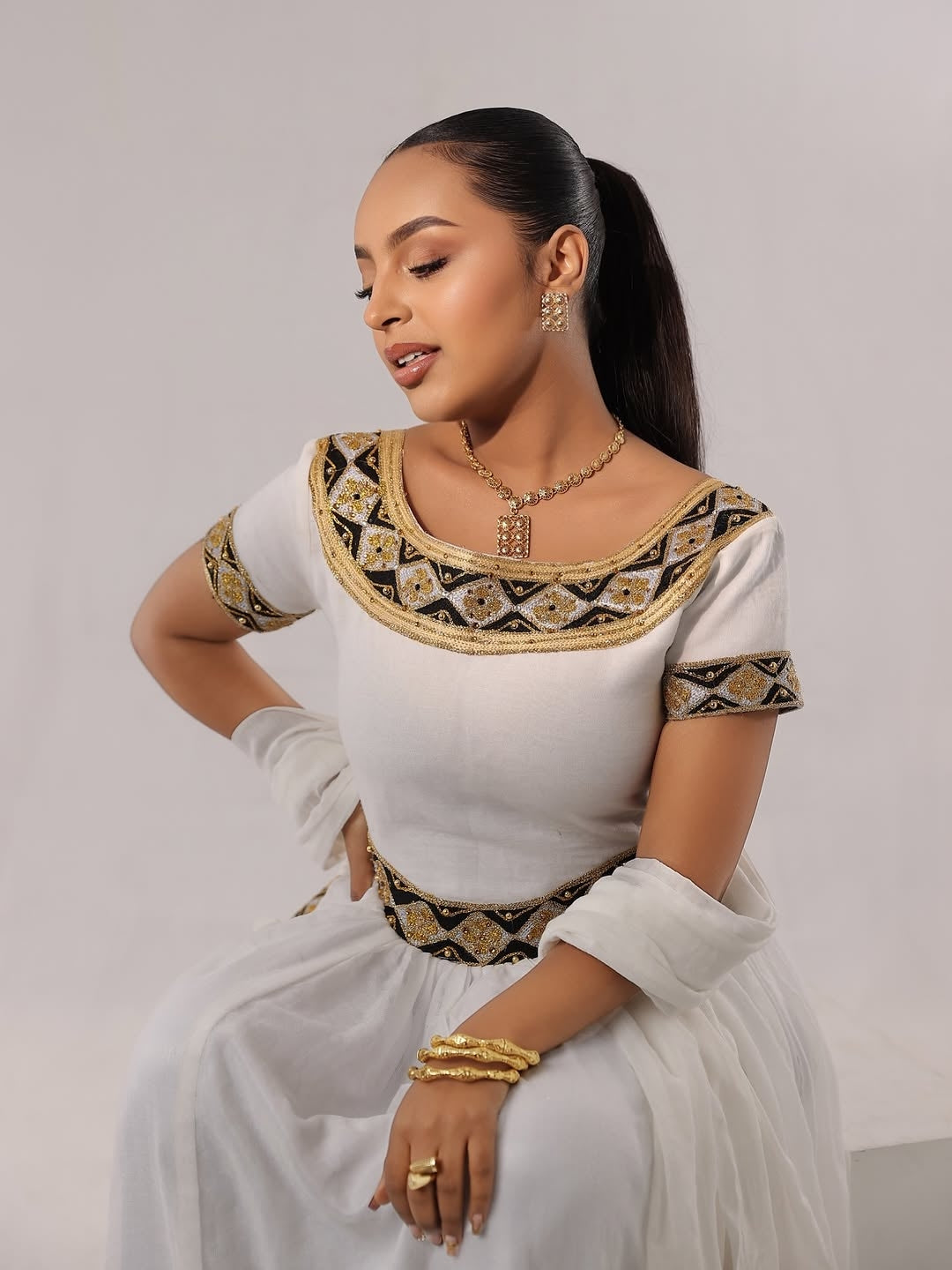 Woman wearing a Habesha Zuria with intricate patterns and gold accents on a plain background