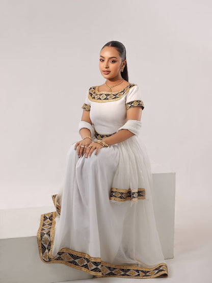 Woman in a white and gold Habesha Zuria sitting on a white stool against a plain background