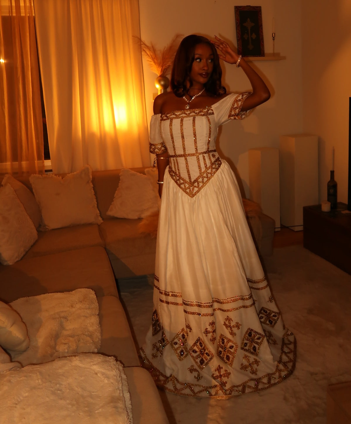 A woman wearing a long off-the-shoulder white Ethiopian Traditional Clothing with ornate gold embroidery and geometric patterns, posing in a warmly lit living room with a sectional sofa and curtains.