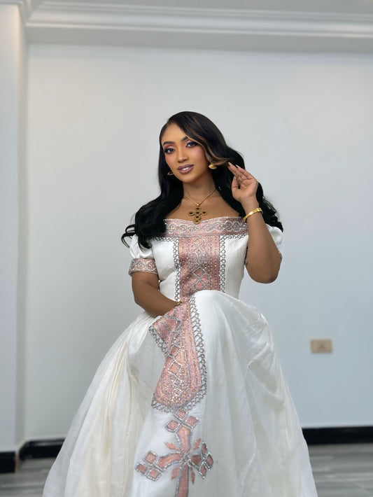 A woman modeling a traditional white Ethiopian Traditional Clothing with pink and silver embroidery, standing in an indoor setting with a plain white wall background.