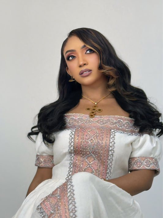A woman with long, wavy dark hair wearing a white traditional Ethiopian Traditional Clothing with pink and silver embroidery, posed against a plain light gray background.