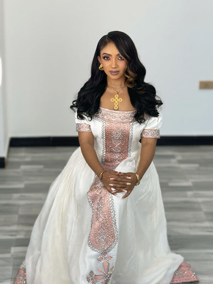 A woman wearing a traditional white Ethiopian Traditional Clothing with pink and silver embroidery, standing in an indoor setting with a gray tiled floor and white walls.