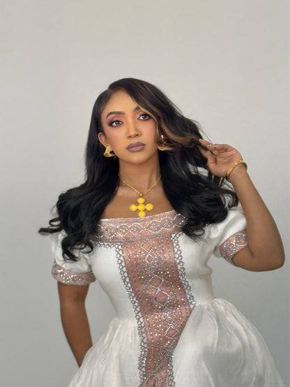 A woman with long, wavy black hair is wearing a white Ethiopian Traditional Clothing with a heavily embellished rose-gold and silver corset-style bodice panel. She is also wearing gold jewelry, including a gold cross pendant, hoop earrings, and a bracelet, against a plain light gray studio background.