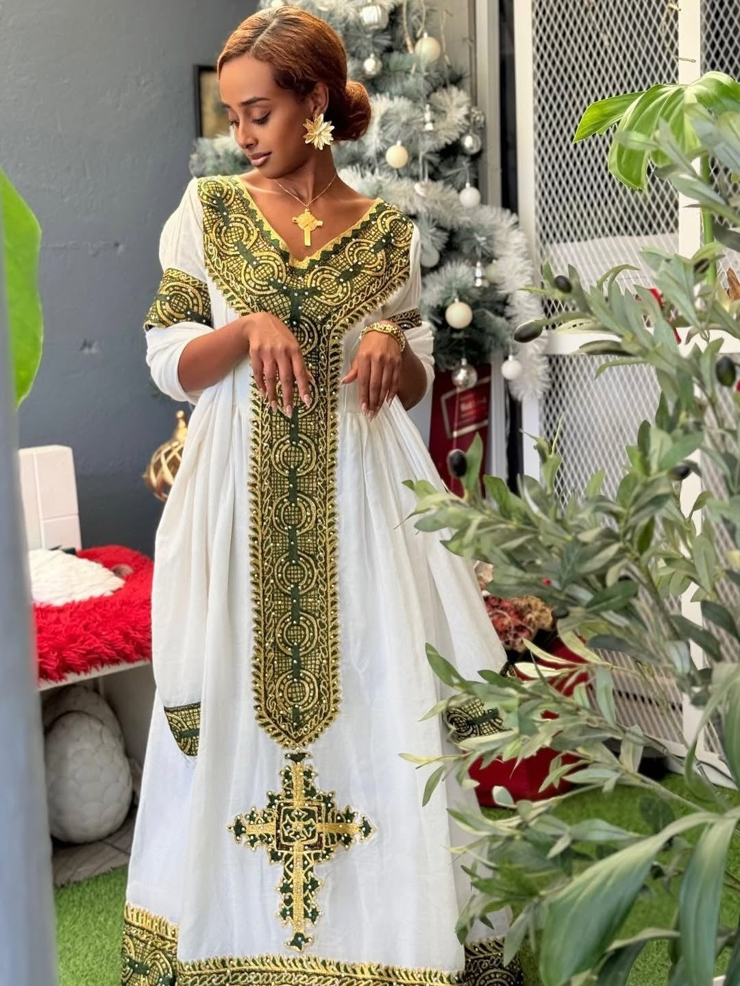 Woman in a white and gold embroidered Habesha Zuria standing in a room with a Christmas tree and plants.