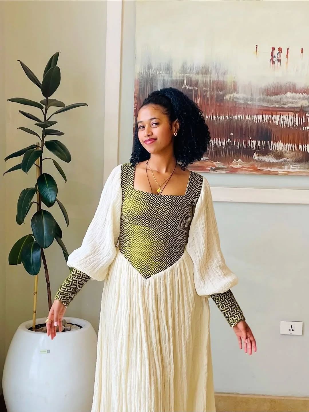 Woman in a long, light-colored habesha dress  standing next to a potted plant indoors.