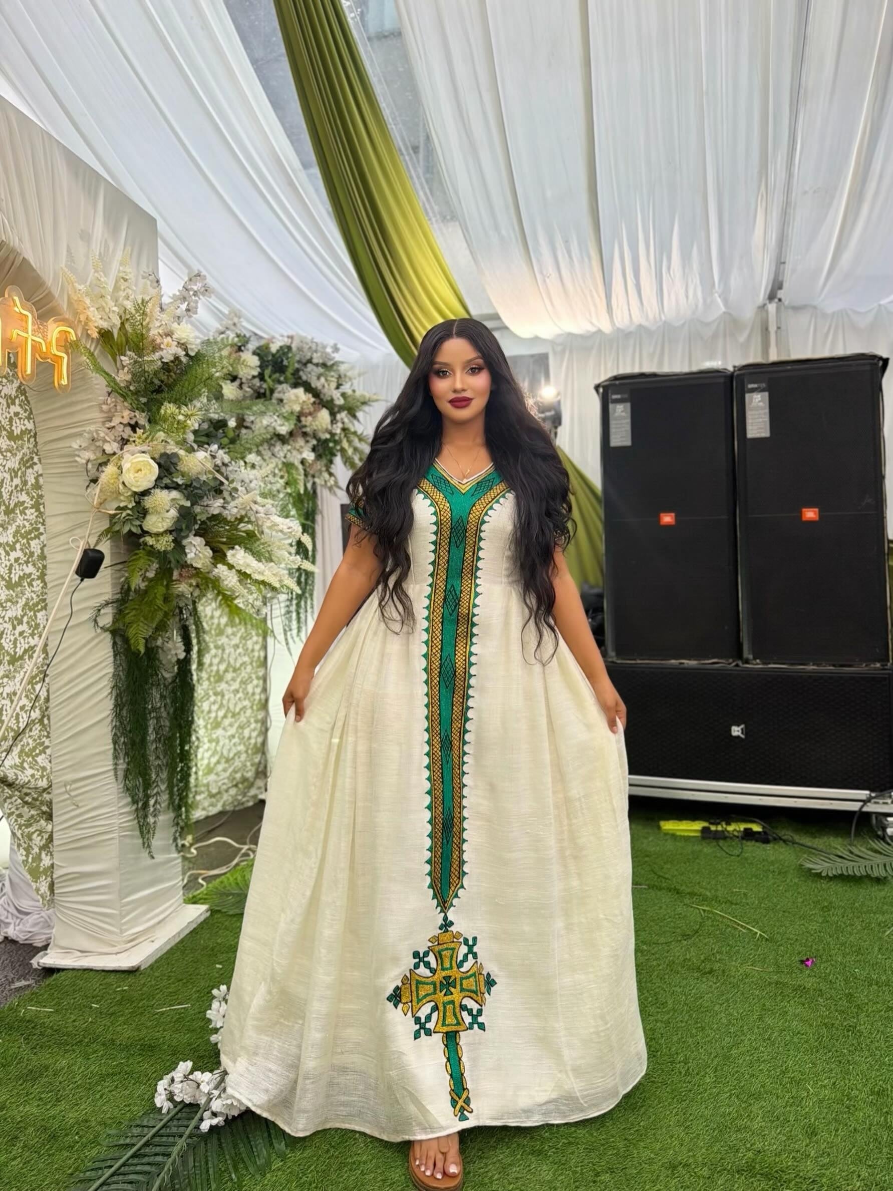 A woman wearing a long white ethiopian dress with green and gold embroidery, standing in an event tent with white drapes, floral arrangements, and speakers in the background.