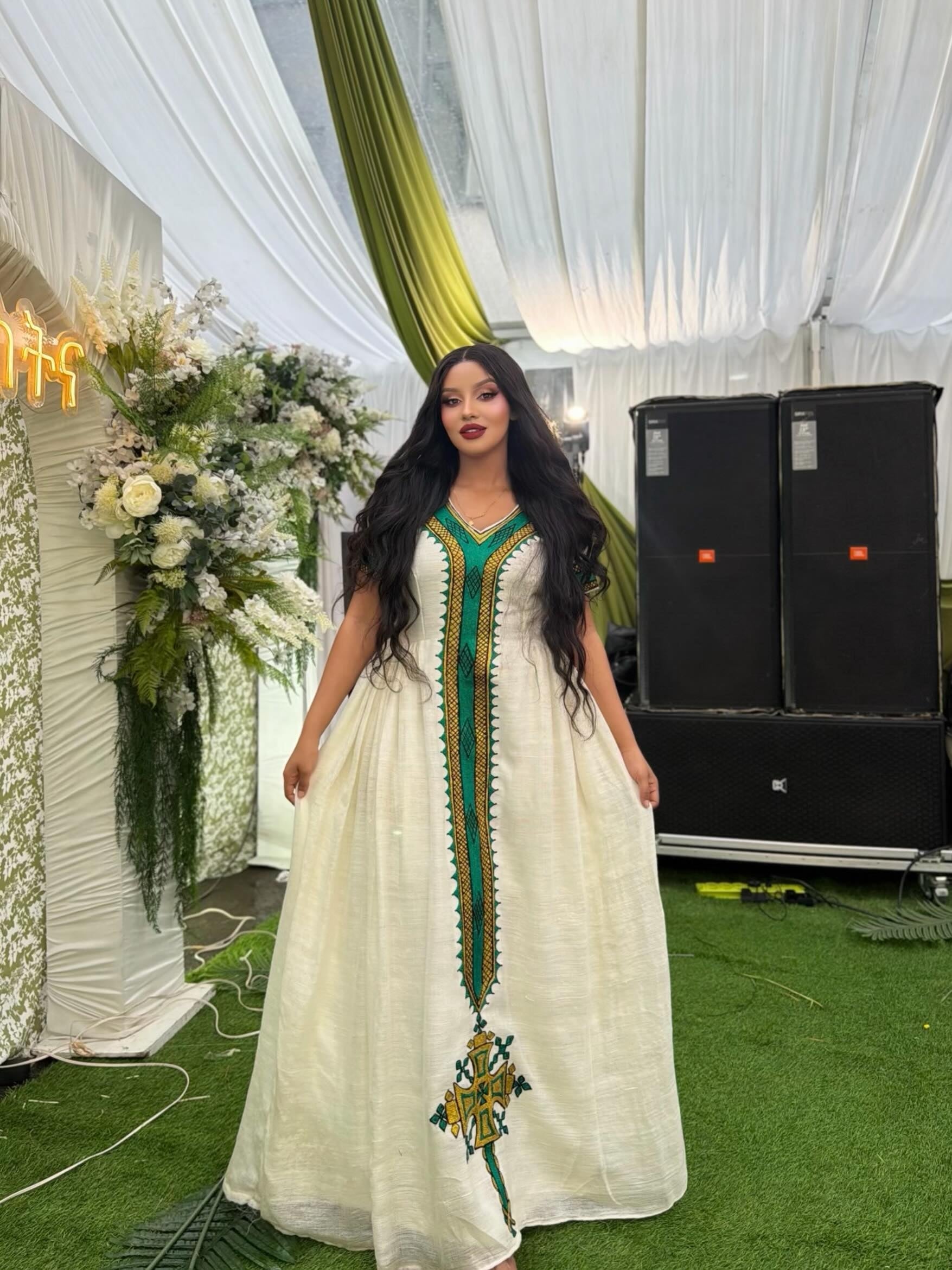 A woman wearing a long white ethiopian dress with green and gold embroidery, standing in front of a decorated event backdrop with speakers and floral arrangements.