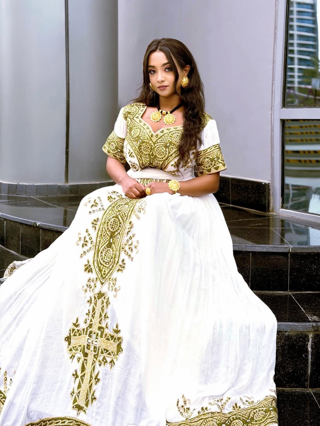 Woman wearing a white and gold embroidered Habesha Dress  sitting on steps.