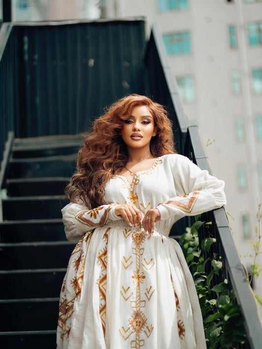 Woman in a white Ethiopian Dress with gold embroidery standing on outdoor steps.