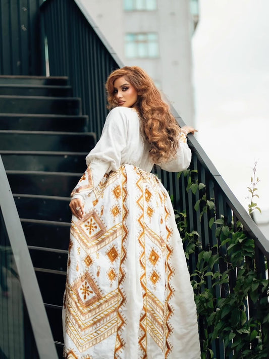 Woman in a long, white Ethiopian Dress with gold patterns standing on a staircase.
