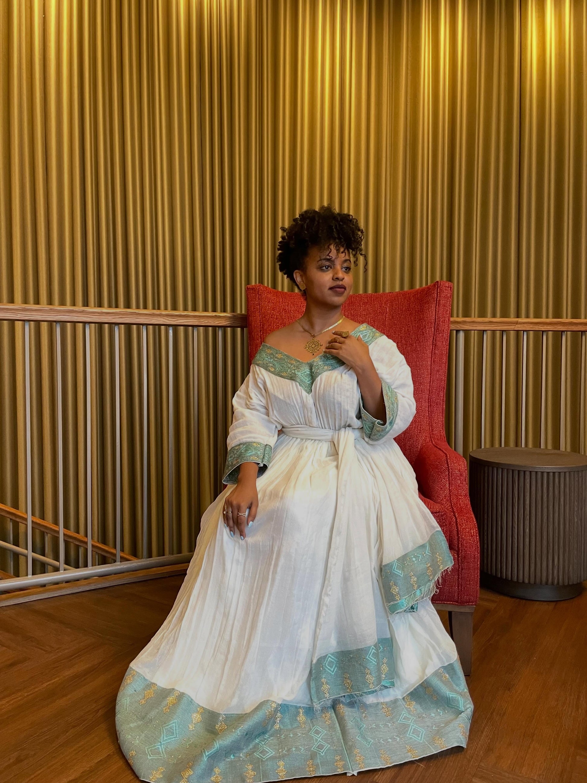 Woman in a white and green dress sitting on a red chair against a gold curtain background