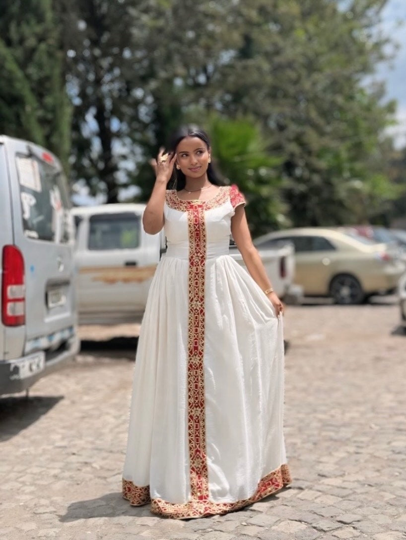 Woman in a white habesha dress with red and gold patterns standing outdoors on a street.