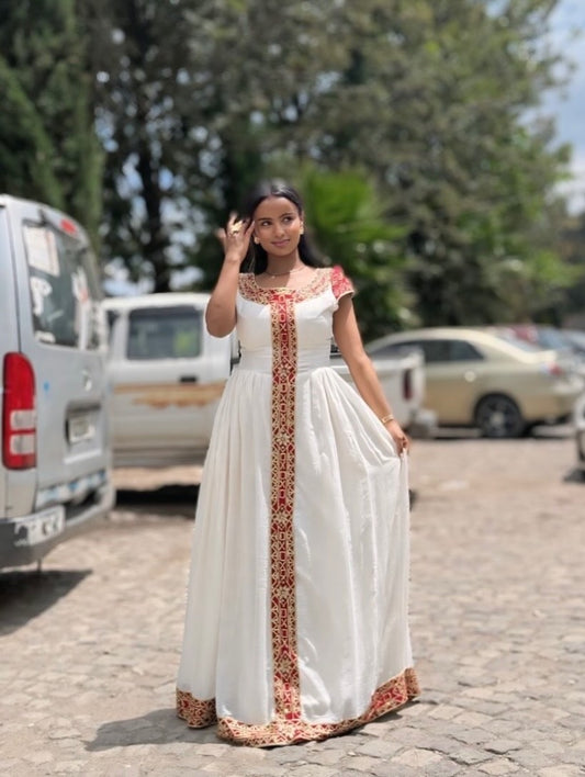 Woman in a white habesha dress with red and gold patterns standing outdoors on a street.