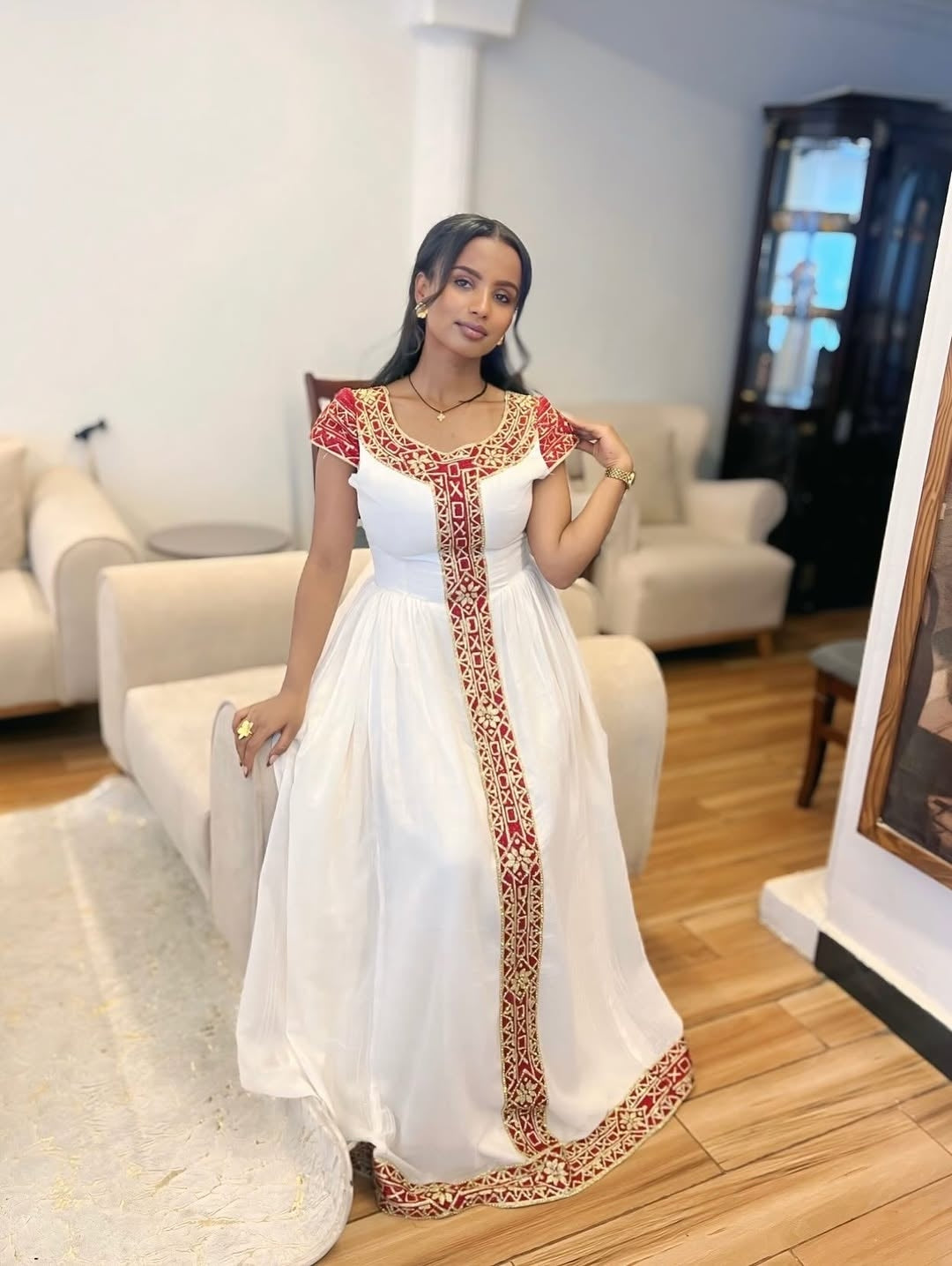 Woman in a white habesha dress with red and gold embroidery in a living room setting