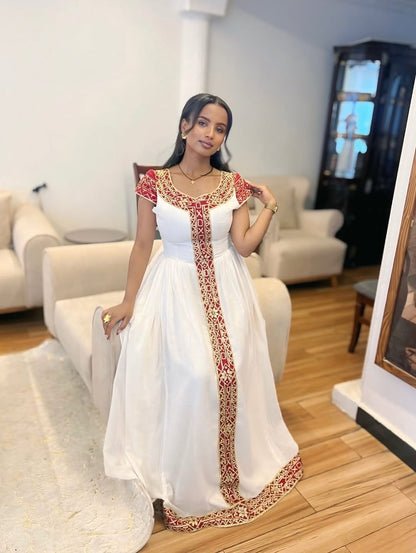Woman in a white habesha dress with red and gold embroidery in a living room setting
