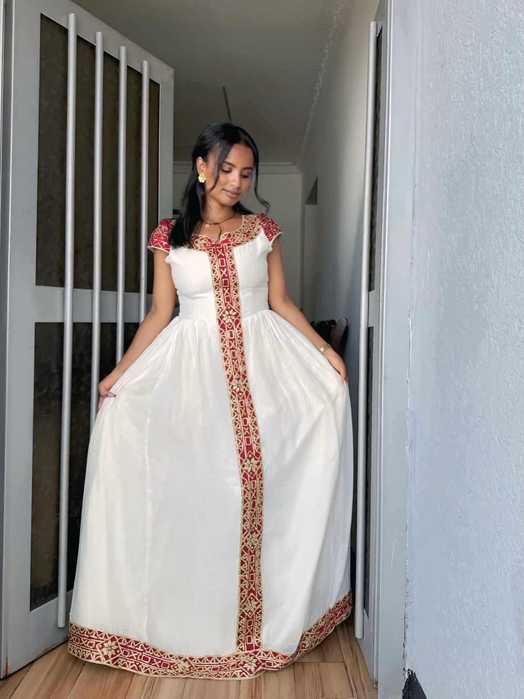 Woman wearing a white habesha dress with red patterns in a room.