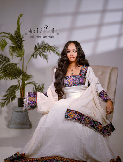A woman seated in a beige armchair wearing a flowing white traditional Ethiopian Dress  with colorful embroidered details on the bodice, sleeves, and hem. A large potted plant stands to the left, and a wall shadow pattern is visible behind her.