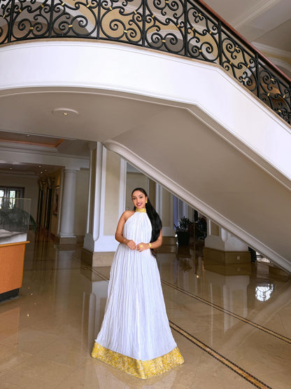 A woman wearing a white sleeveless pleated Ethiopian shop near me  with a gold collar and a gold patterned hem stands in a grand indoor lobby with a staircase and polished floor.