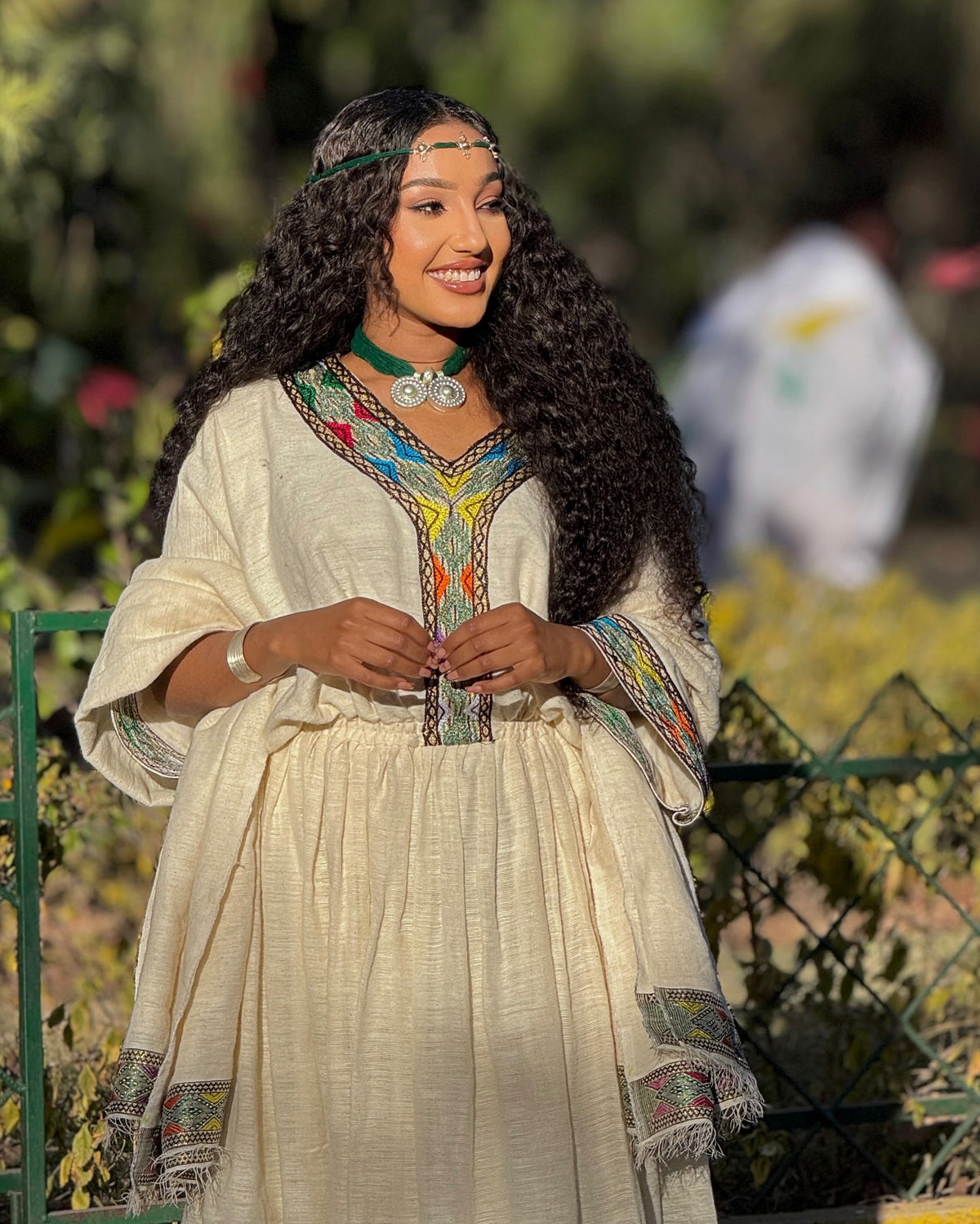 A woman with long, curly black hair wearing a cream-colored traditional Ethiopian Clothing with colorful embroidered details, standing outdoors in front of a green fence.