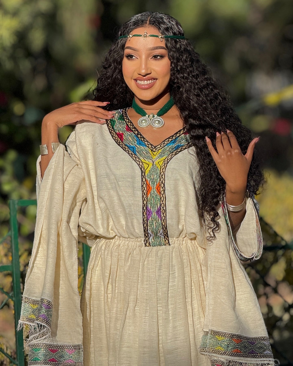 A woman wearing a cream-colored bohemian-style Ethiopian Clothing with colorful embroidered details on the neckline and cuffs, posing outdoors with a green garden background.