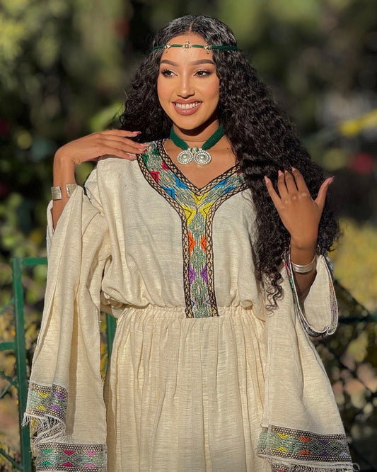 A woman wearing a cream-colored bohemian-style Ethiopian Clothing with colorful embroidered details on the neckline and cuffs, posing outdoors with a green garden background.