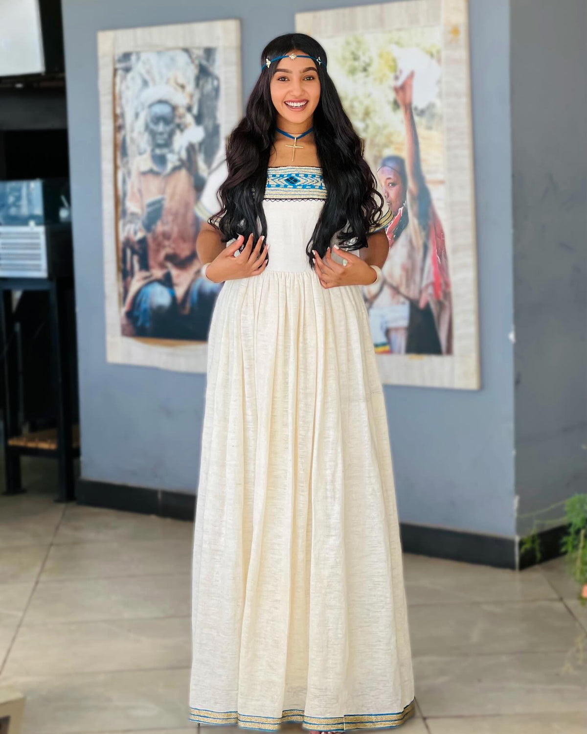 A woman wearing a long cream-colored linen Ethiopian National Dresses with blue and gold embroidery on the bodice and hem, standing in an indoor setting with framed art on a gray wall.