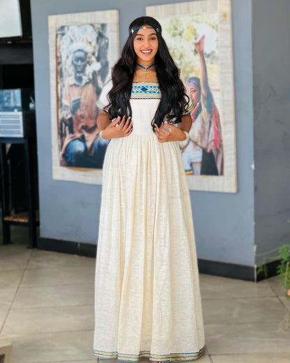 A woman wearing a long cream-colored linen Ethiopian National Dresses with blue and gold embroidery on the bodice and hem, standing in an indoor setting with framed art on a gray wall.