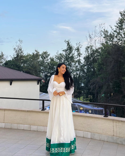 A woman wearing a white Ethiopian Clothing  with a green patterned hem and sheer white sleeves, standing on an outdoor balcony with trees and a building in the background.