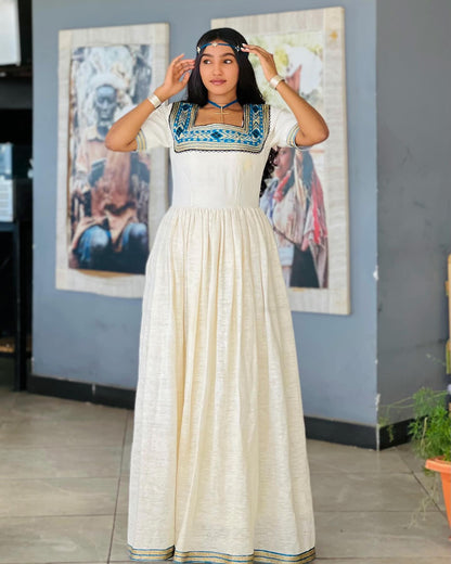 A woman modeling a cream-colored, floor-length Ethiopian National Dresses with a square neckline decorated with blue and green embroidery, standing in an indoor setting with framed art on a gray wall.