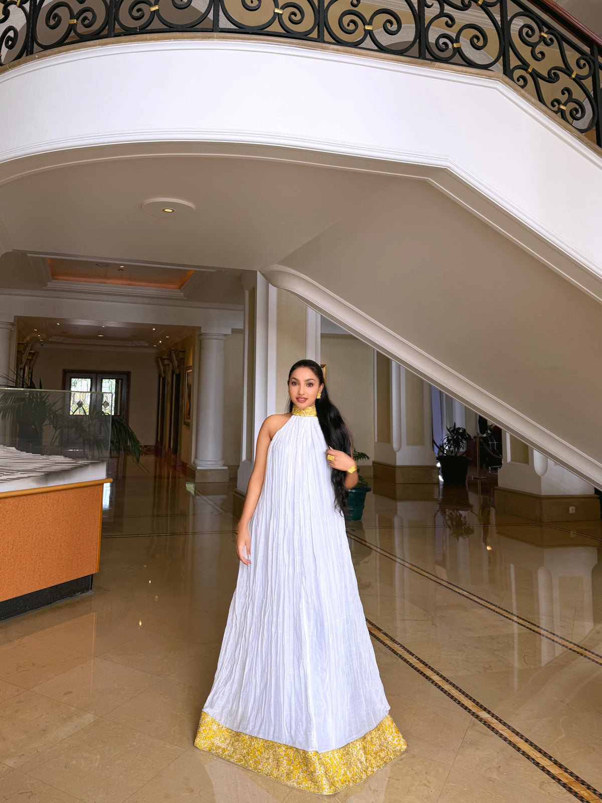 A woman wearing a white sleeveless pleated Ethiopian shop near me  with a gold embroidered hem and a gold collar, standing in a grand, upscale indoor lobby with a staircase and polished floors.