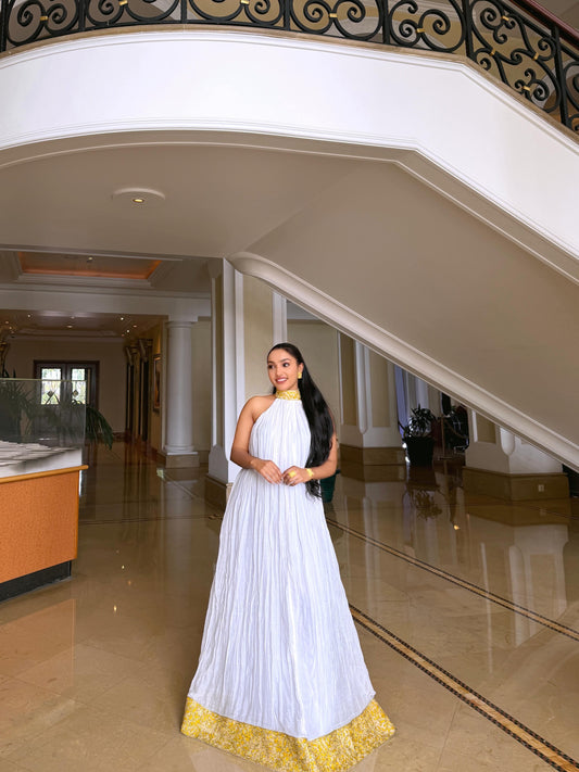 A woman wearing a white halter-neck maxi Ethiopian shop near me  with a gold embroidered hem stands in a grand hotel lobby with a staircase in the background.