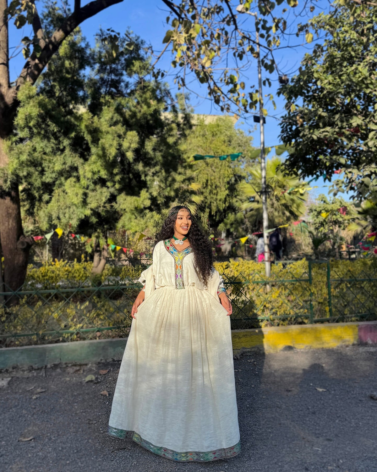 A woman wearing a long white traditional Ethiopian Clothing with colorful embroidery stands outdoors in a park with trees, greenery, and a street sign in the background.