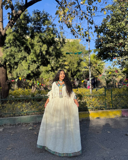 A woman wearing a long white traditional Ethiopian Clothing with colorful embroidery stands outdoors in a park with trees, greenery, and a street sign in the background.