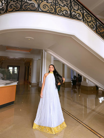 A woman wearing a white sleeveless pleated Ethiopian shop near me  with a gold patterned hem stands in a grand indoor lobby with a staircase and polished floor.