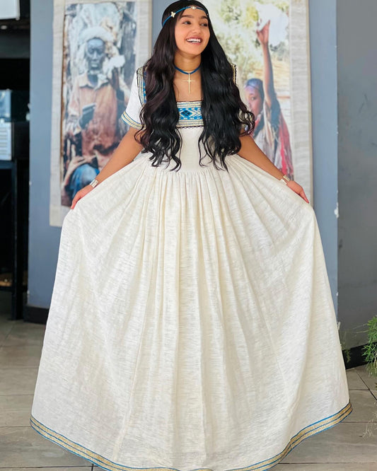 A woman wearing a traditional Indian Ethiopian National Dresses with a wide, pleated off-white skirt and a short embroidered blouse, holding out the sides of the skirt to show its volume. She is standing indoors in front of a gray wall with framed art.
