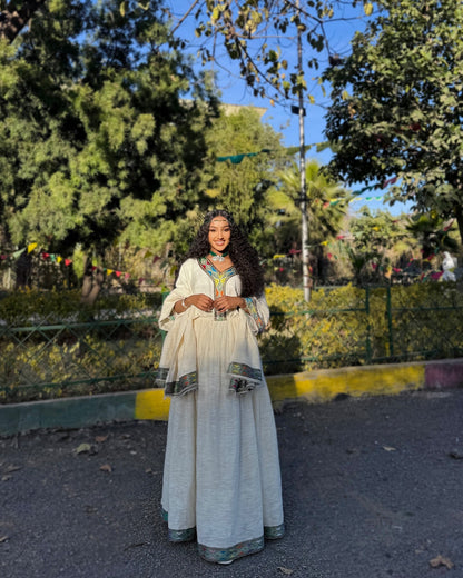 A woman wearing a traditional long white Ethiopian Clothing with green and gold embroidery and a matching headpiece, standing outdoors in a park-like setting with trees, a fence, and a blue sky in the background.