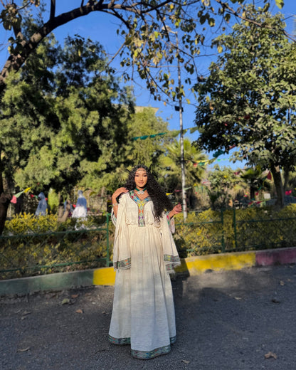 A woman wearing a long cream-colored traditional Ethiopian Clothing with colorful embroidered details stands outdoors in a park setting with trees, blue sky, and a fence in the background.