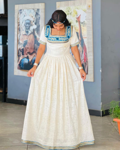 A woman wearing a long cream-colored linen Ethiopian National Dresses with blue and green geometric embroidery on the bodice and sleeves, standing in an indoor setting with framed art on a gray wall.