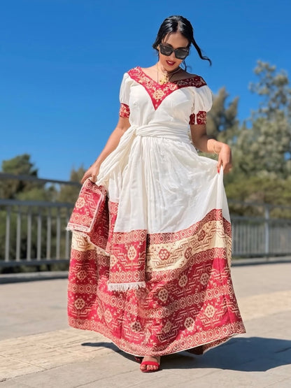 Woman wearing a white and red Ethiopian cultural dress with a patterned design outdoors on a sunny day.
